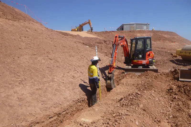 Surveyor and mini excavator cutting a trench on a batter