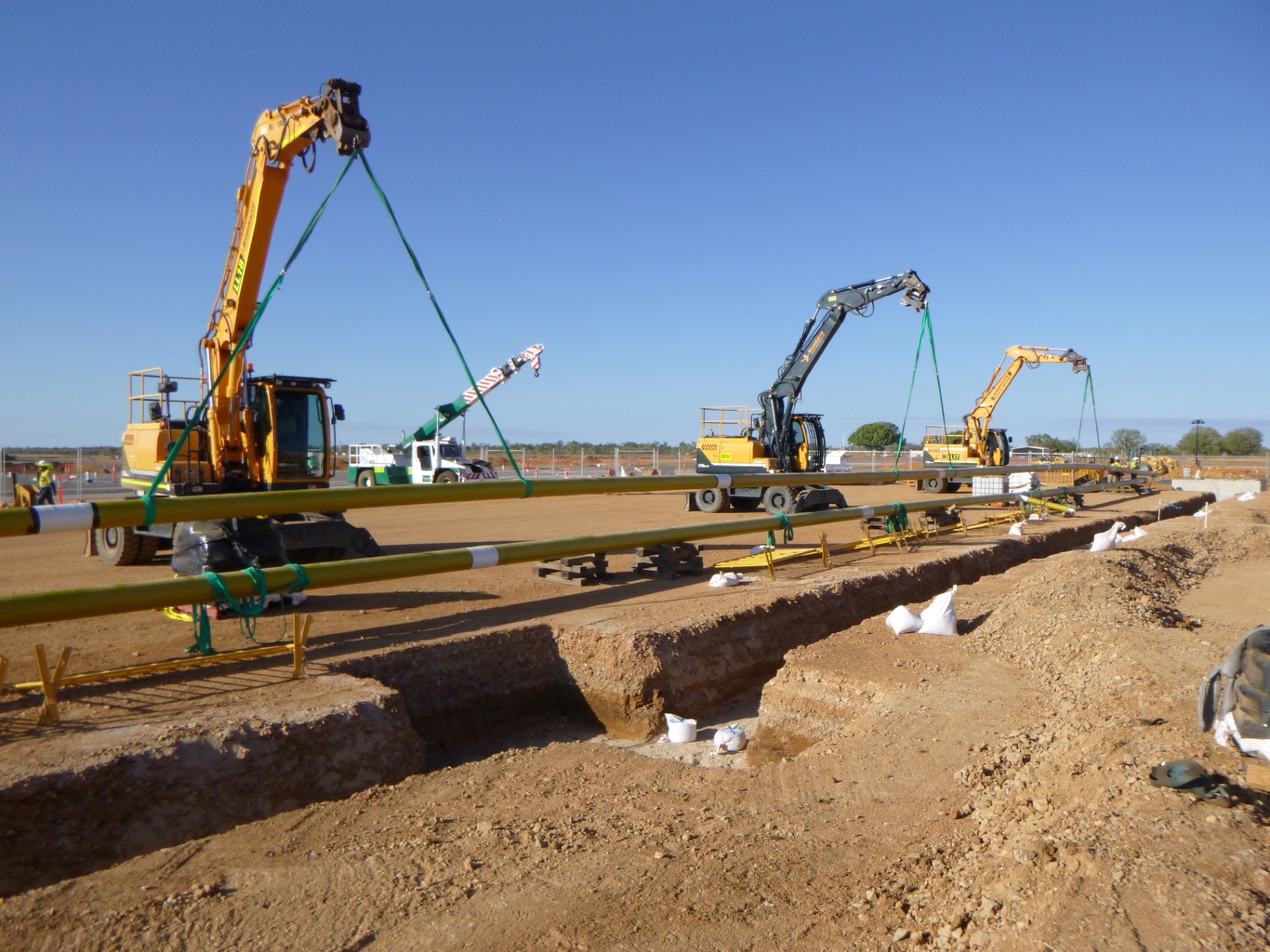 Three excavators lifting gas pipeline sections in unison beside open trench